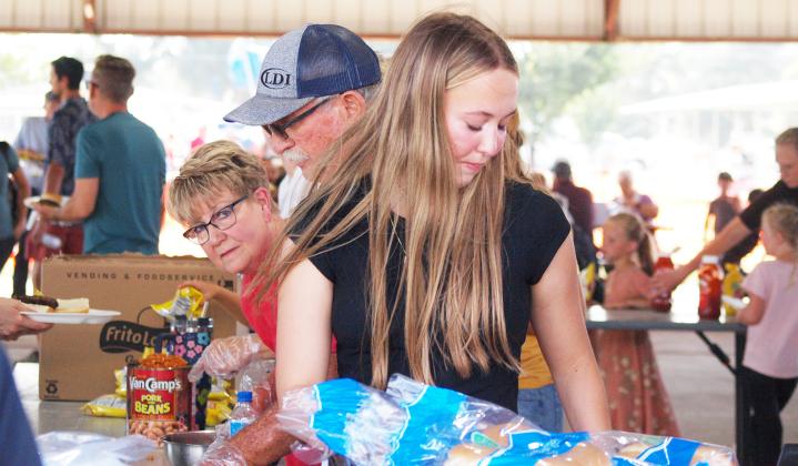 Volunteer Christy Marcy peeks around her husband, Mike, and granddaughter, Audrina, at the Free Bar-B-Q and Watermelon Feed on Thursday, August 1.